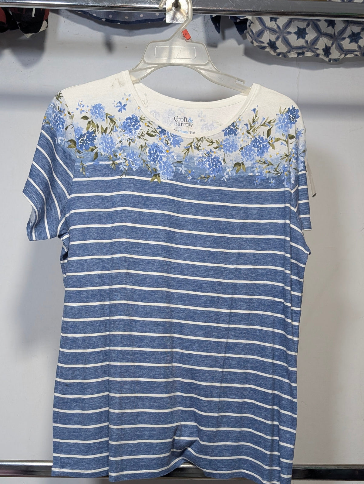 A short-sleeve crewneck t-shirt hanging on a clear plastic hanger. The shirt features a blue and white horizontal stripe pattern across the body, with a white yoke at the shoulders decorated with a blue and green floral print.