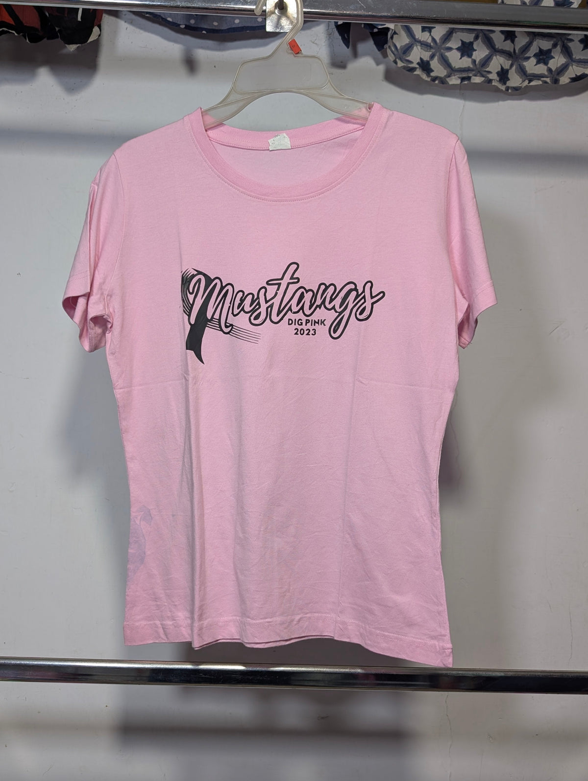 A light pink short-sleeve t-shirt hanging on a hanger, featuring a black graphic print on the front that reads 'Mustangs 2025' with a horse silhouette. The shirt is photographed against a plain wall in what appears to be a storage or retail space.