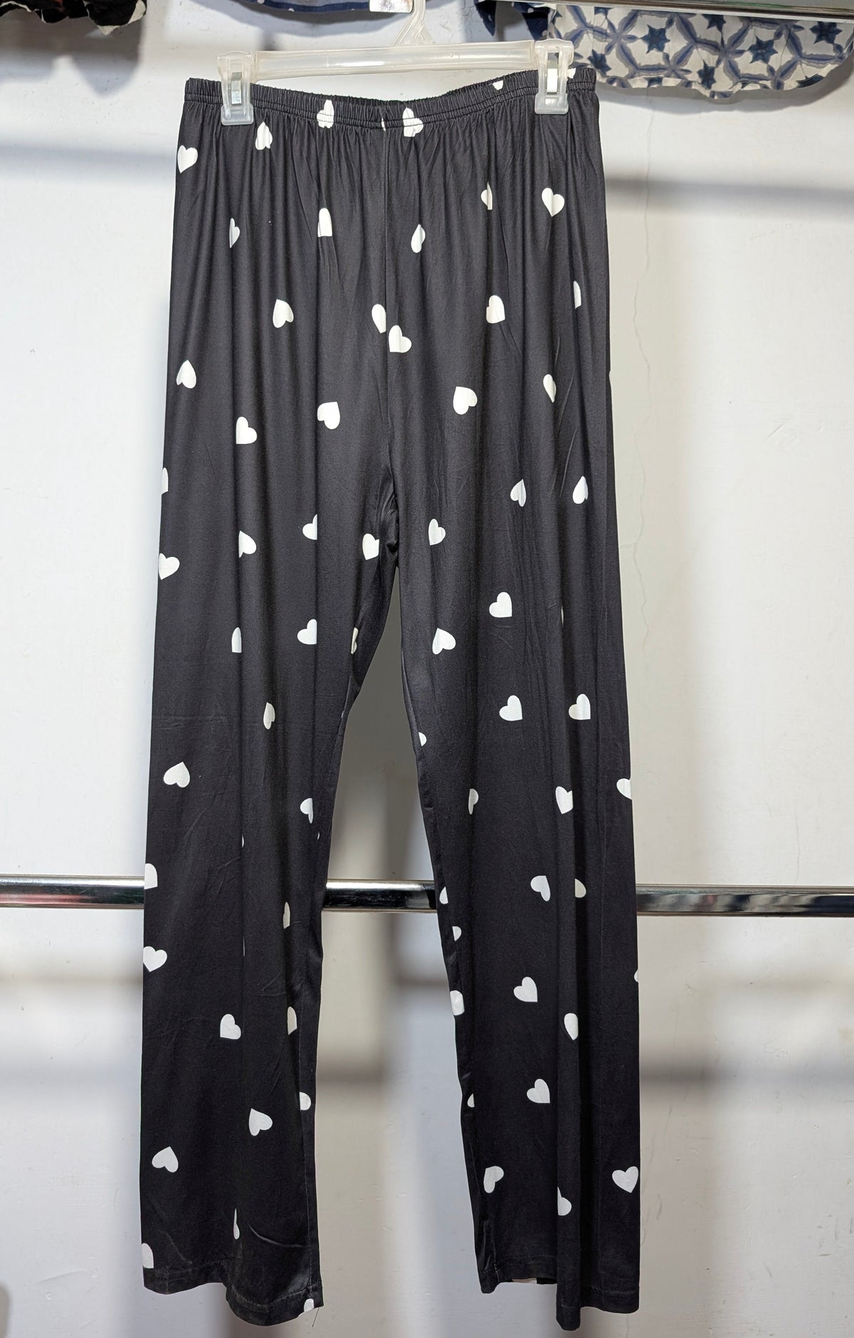 Black cropped pants with a white heart pattern, hanging on a white hanger from a metal clothing rack.