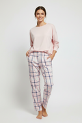 A woman modeling a light pink long-sleeve crewneck top and matching pink pajama pants with a navy blue windowpane plaid pattern, standing against a light gray studio background.
