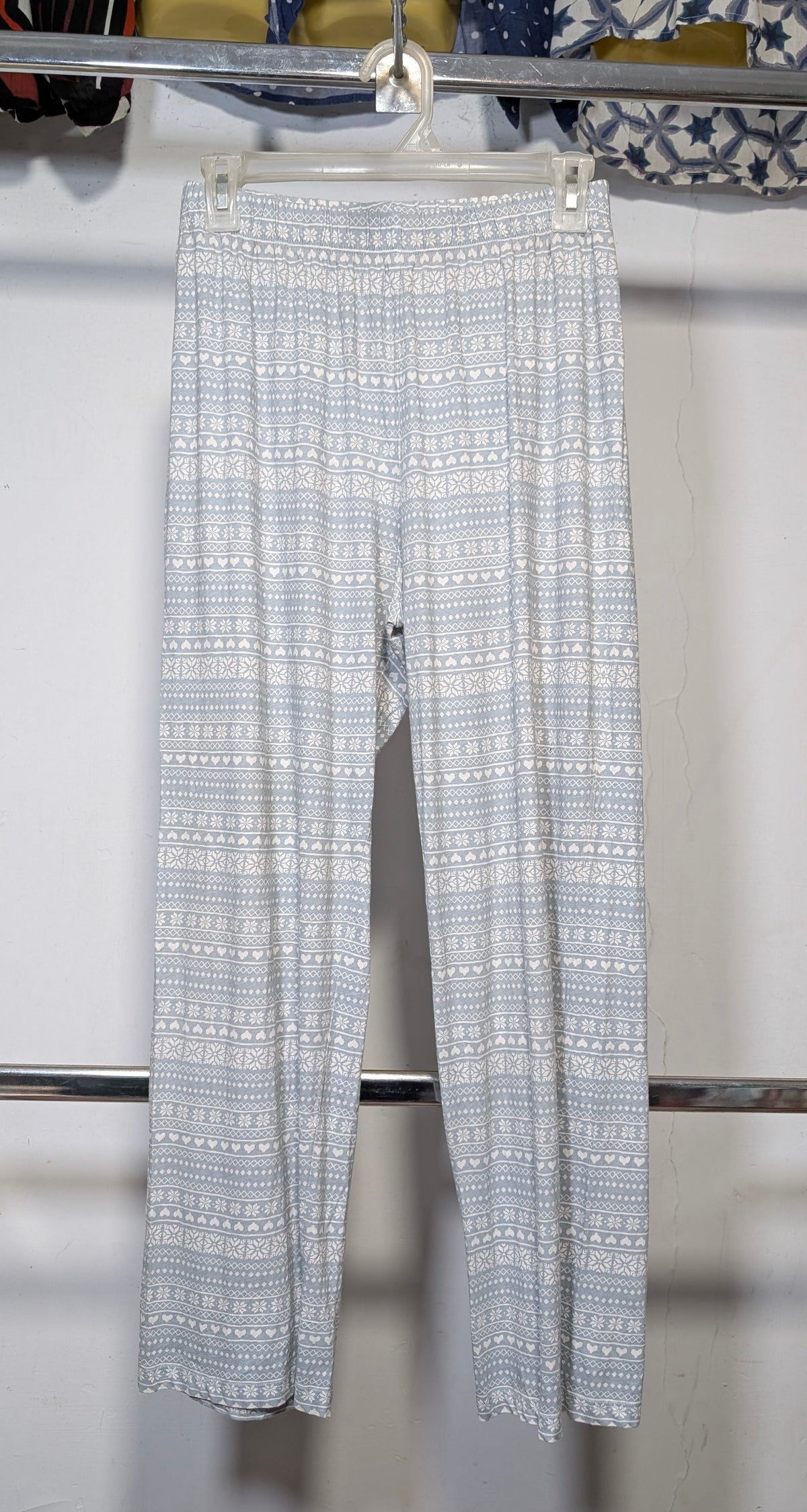Light blue and white patterned wide-leg pants hanging on a hanger from a clothing rack, set against a white wall with other garments visible in the background.