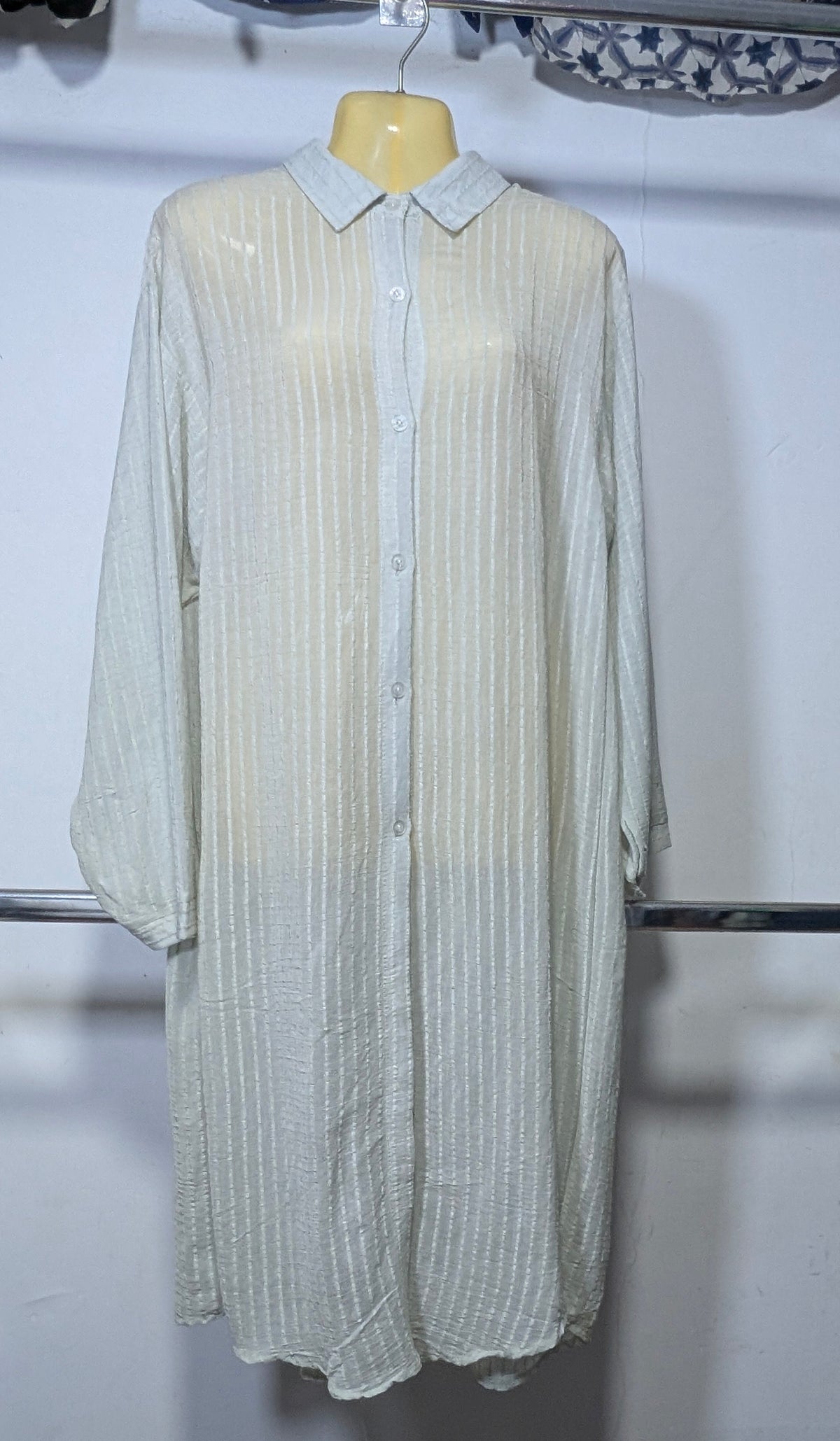 A long-sleeve button-up shirt dress in a light cream or off-white color with a subtle vertical textured stripe pattern, displayed on a mannequin torso against a white wall.