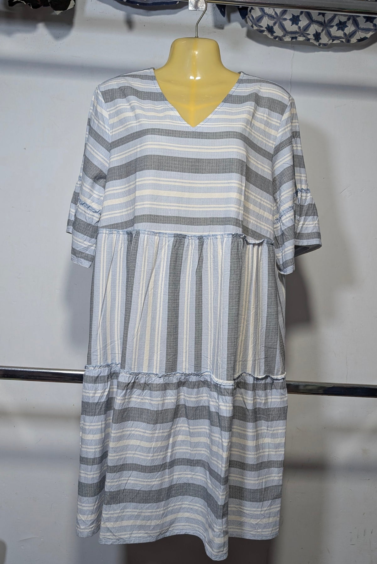 A tiered short-sleeve dress with gray, white, and light blue horizontal stripes, displayed on a yellow mannequin hanger on a clothing rack.