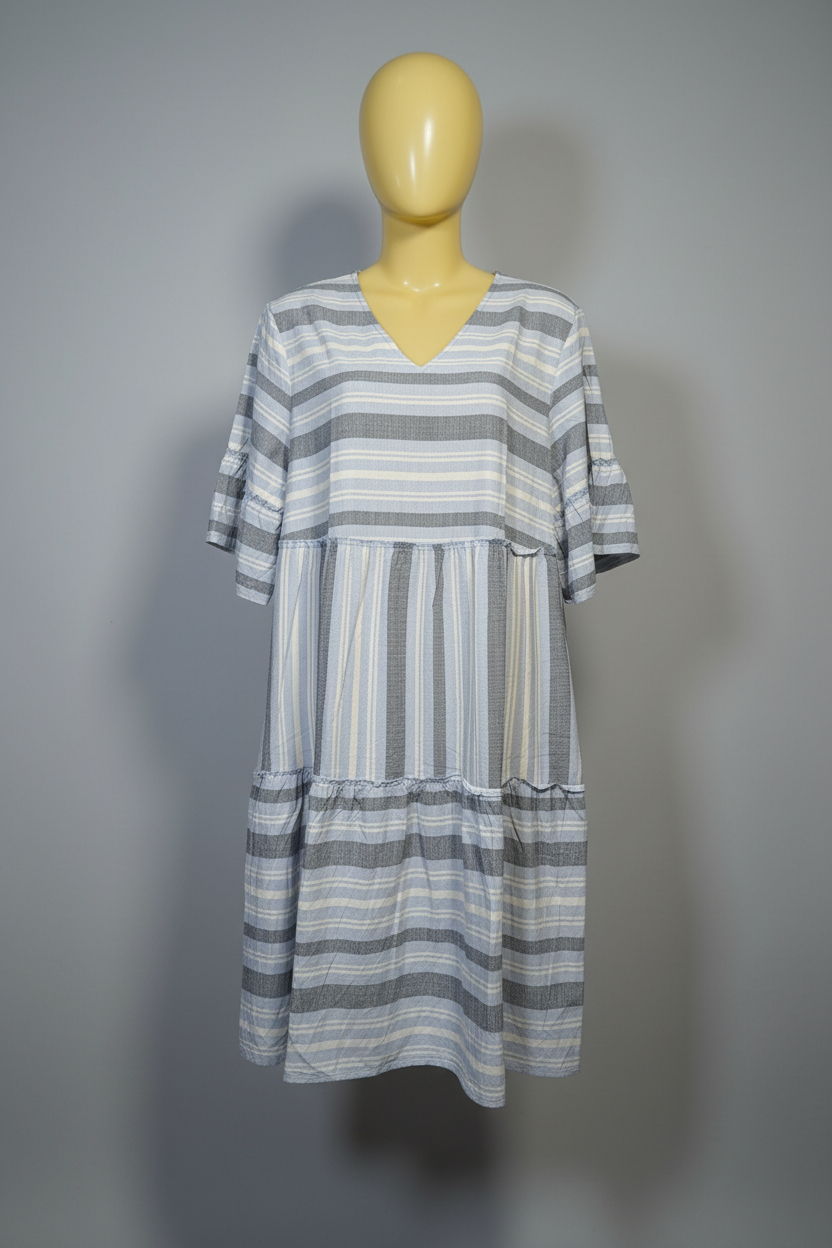 A woman modeling a tiered, short-sleeve V-neck dress with gray, white, and light blue vertical stripes, paired with light-wash blue jeans and white sneakers, against a solid beige studio background.