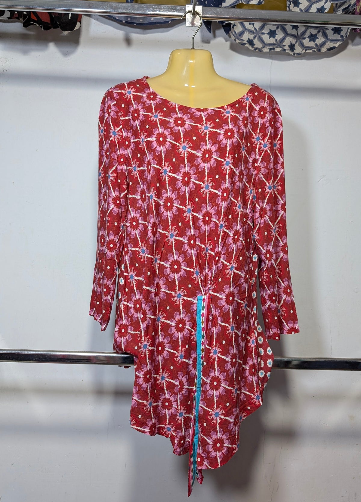A red and white patterned long-sleeve tunic top with a tie-dye style floral design, hanging on a yellow mannequin torso on a clothing rack.