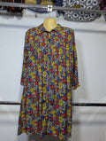 A long-sleeve button-up shirt dress with a dense multicolor floral print, hanging on a wooden hanger against a white wall.
