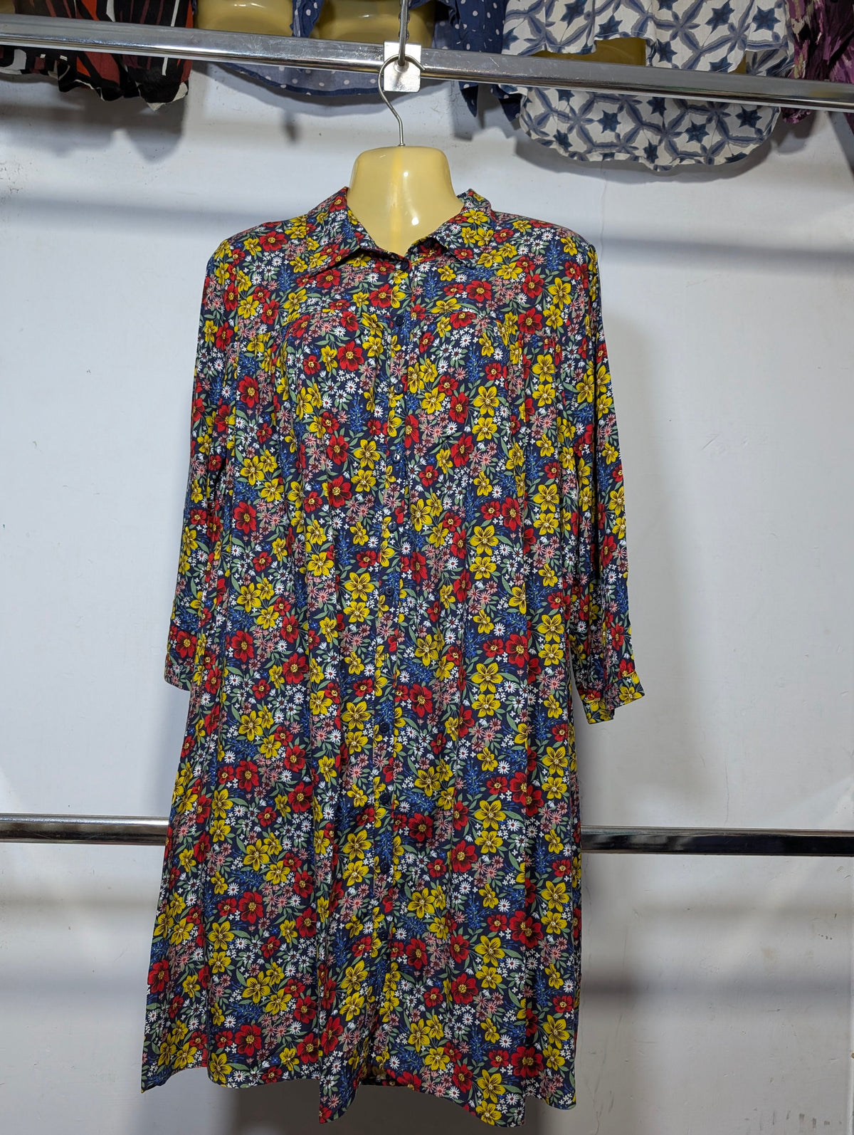 A long-sleeve button-up shirt dress with a dense multicolor floral print, hanging on a wooden hanger against a white wall.