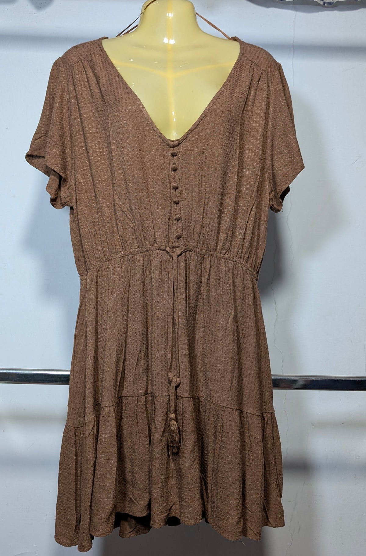 A brown short-sleeve dress with a V-neckline and button details, displayed on a mannequin torso against a white wall.