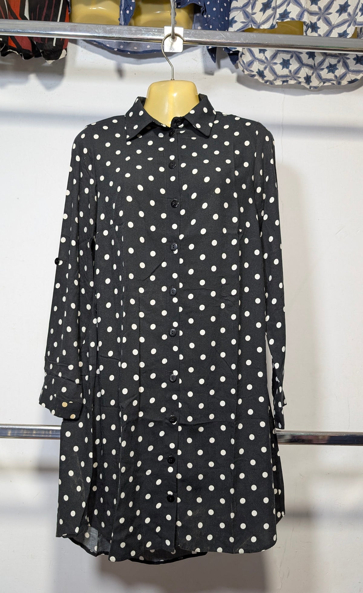 A black long-sleeve button-up shirt with white polka dots, displayed on a yellow hanger on a clothing rack.