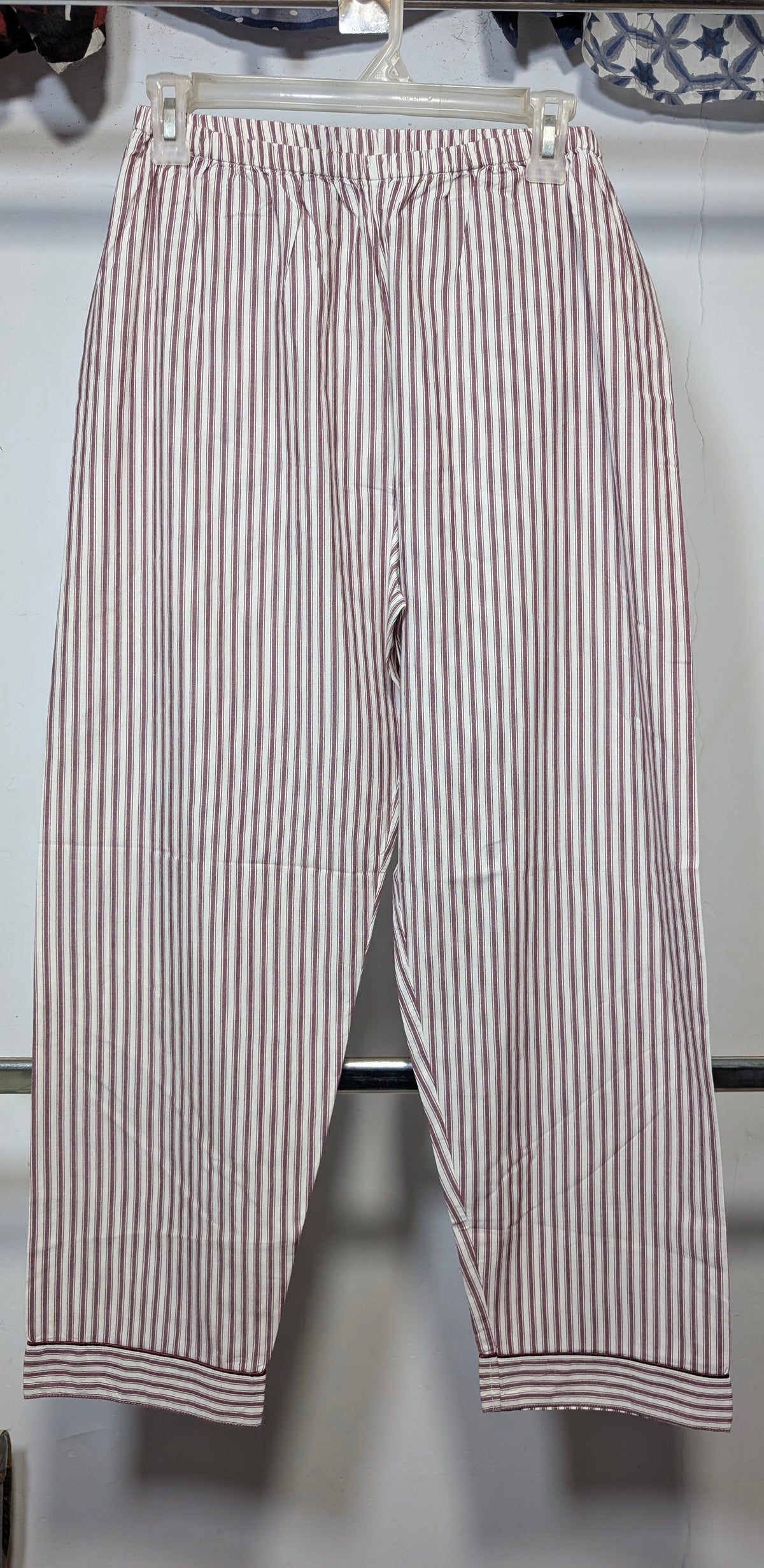 A pair of cropped pants with vertical red and white stripes, hanging on a clear plastic hanger against a white wall.