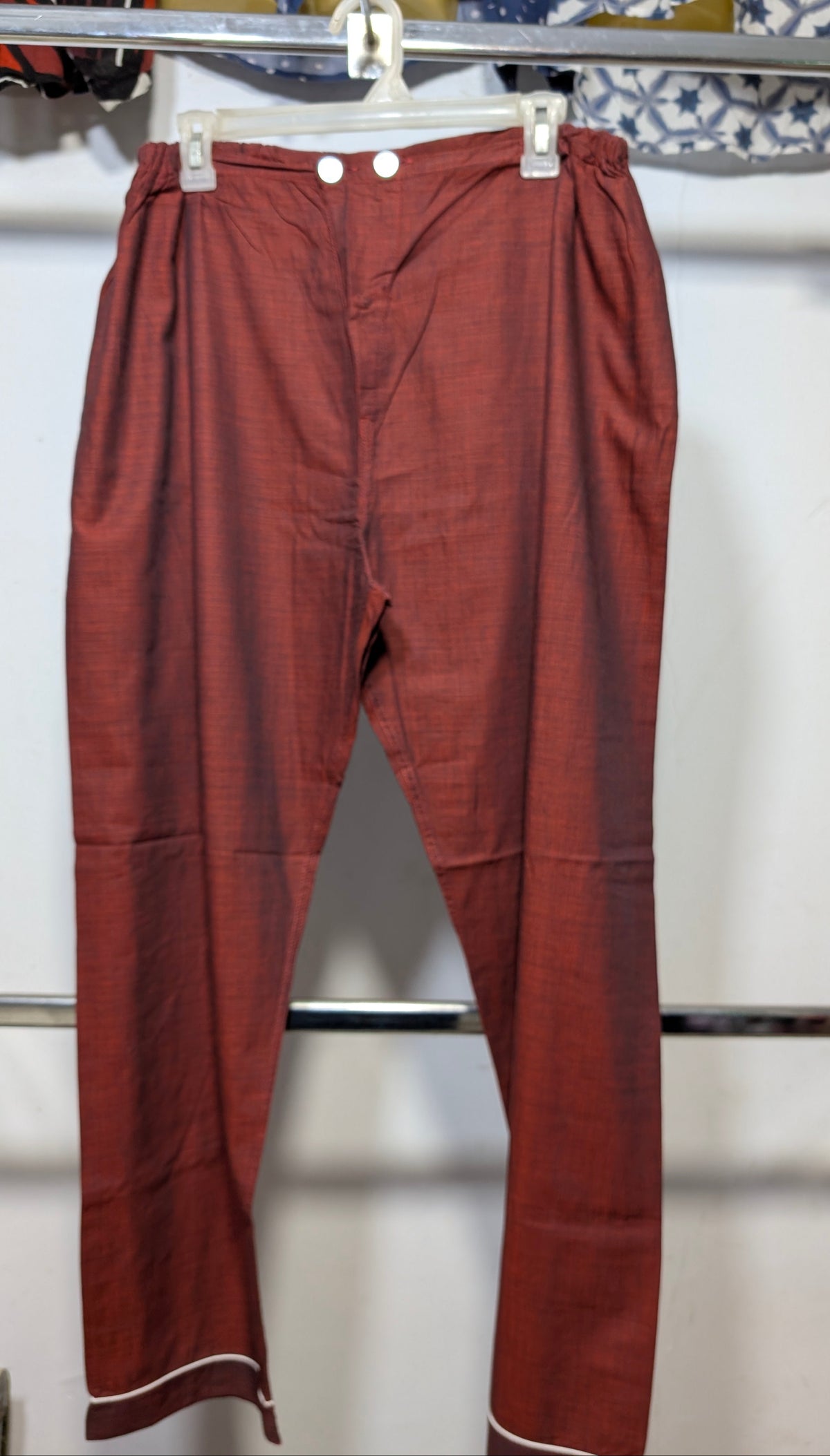 A pair of rust-red linen trousers hanging on a white plastic hanger from a clothing rack, featuring a relaxed straight-leg fit and white piping along the hem.