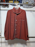 A men's long-sleeve button-up pajama shirt in a rust red or terracotta color, featuring white piping along the collar, front placket, and chest pocket, hanging on a white hanger against a white wall.