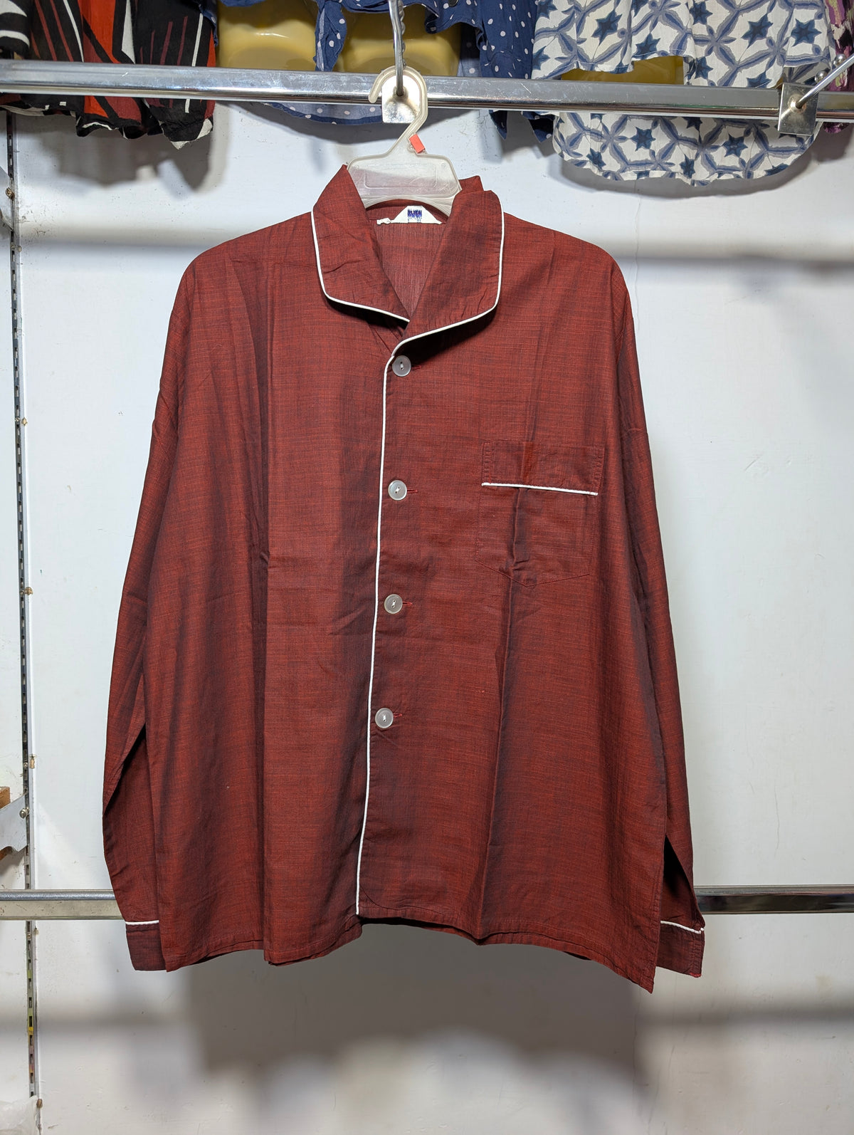 A men's long-sleeve button-up pajama shirt in a rust red or terracotta color, featuring white piping along the collar, front placket, and chest pocket, hanging on a white hanger against a white wall.