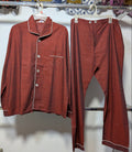 A two-piece pajama set consisting of a long-sleeve button-front top and matching pants, both in a rust-red or terracotta color with thin white piping trim, hanging on white plastic hangers on a clothing rack.