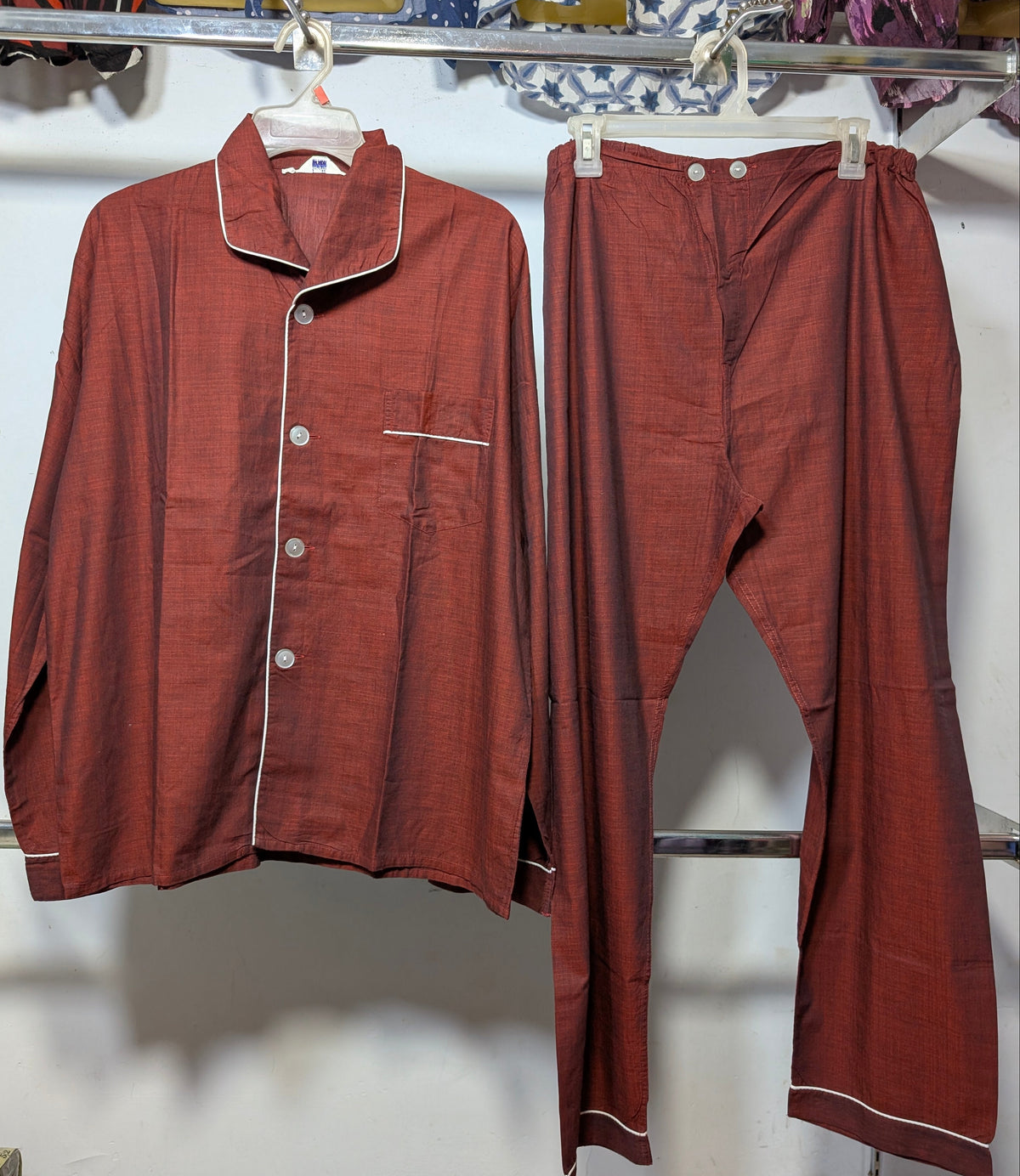 A two-piece pajama set consisting of a long-sleeve button-front top and matching pants, both in a rust-red or terracotta color with thin white piping trim, hanging on white plastic hangers on a clothing rack.