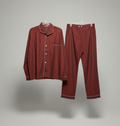 A two-piece pajama set in a muted brick red color, featuring a long-sleeve button-up shirt with contrasting white piping and matching trousers, hanging on white hangers against a neutral gray background.