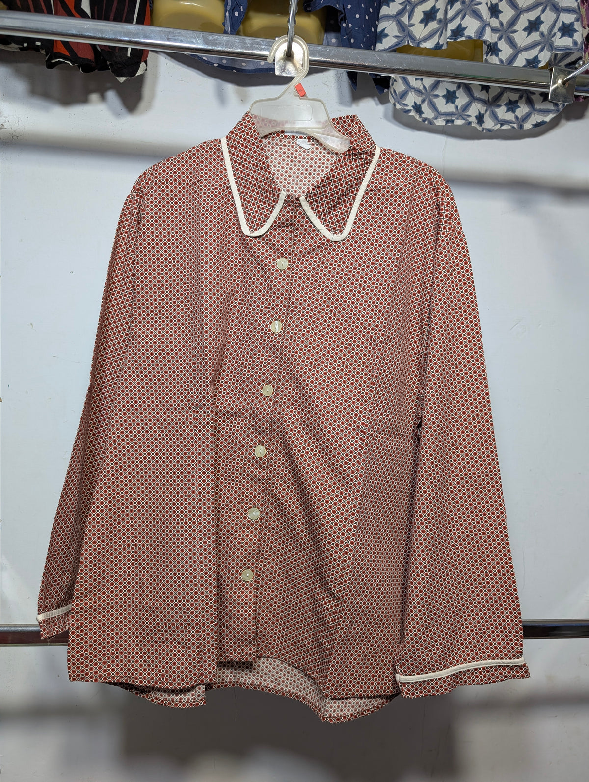 A long-sleeve button-up shirt with a small red and white polka dot pattern, featuring a white Peter Pan collar and white piping along the edges, hanging on a white hanger.