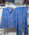 A two-piece pajama set consisting of a long-sleeve button-up shirt and matching pants, both in a medium blue fabric with a colorful floral print. The shirt is shown on the left hanging on a white hanger, and the pants are on the right, also on hangers, against a white wall.