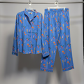 A two-piece pajama set hanging on white hangers from a metal rail. The set includes a long-sleeve button-up shirt and matching pants in a medium blue fabric with an allover print of orange flowers, green stems, and small yellow accents.