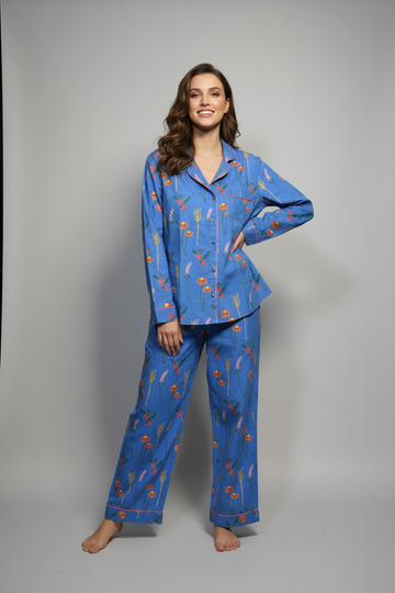 A woman modeling a two-piece pajama set with a blue base and a pattern of orange flowers and green leaves, standing against a solid gray studio background.