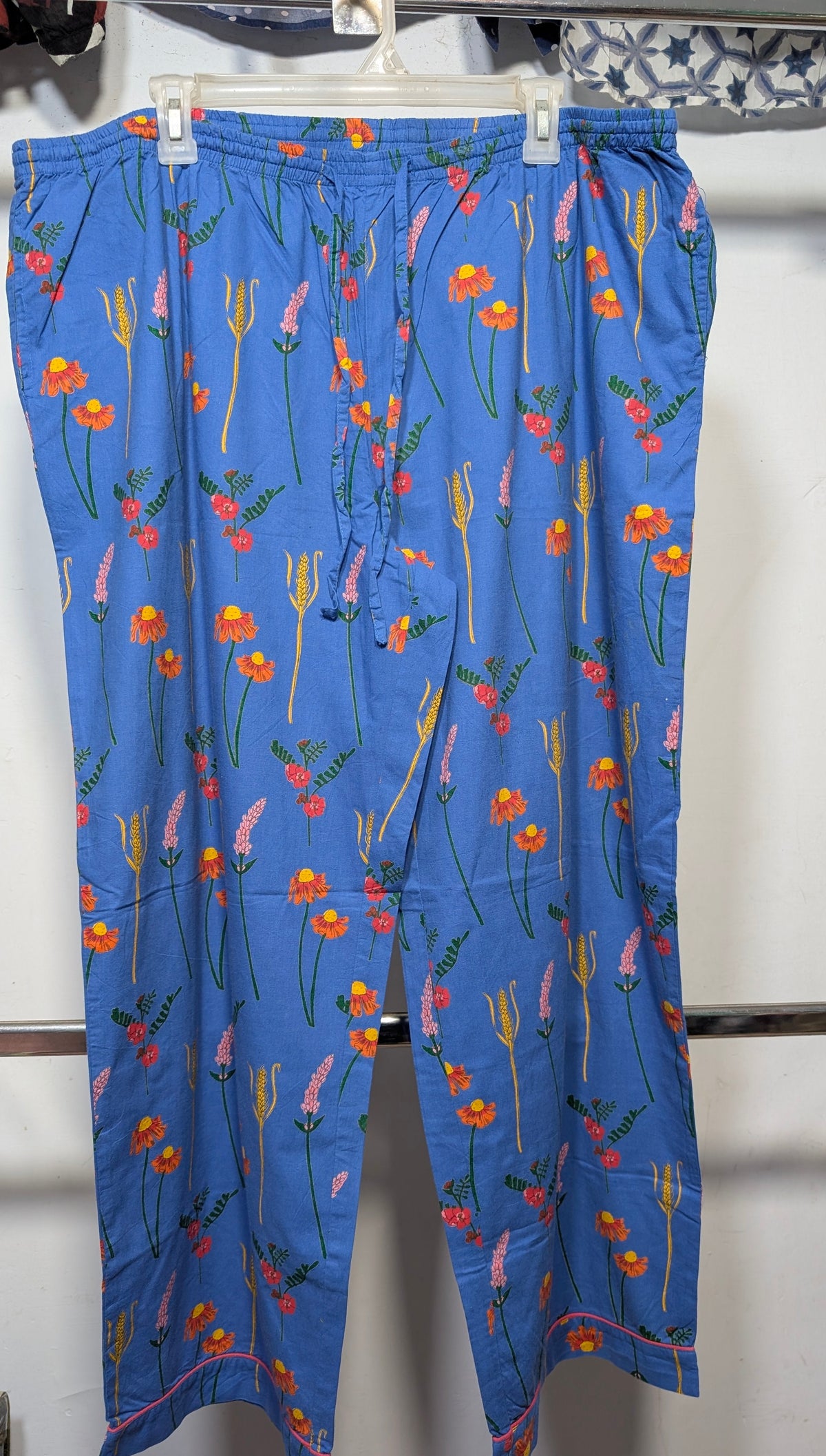 Blue cropped pants with a colorful floral and botanical print, hanging on a white plastic hanger against a white wall.