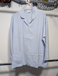 A light blue long-sleeve pajama shirt with white piping trim and gold buttons, hanging on a clear plastic hanger against a white wall.