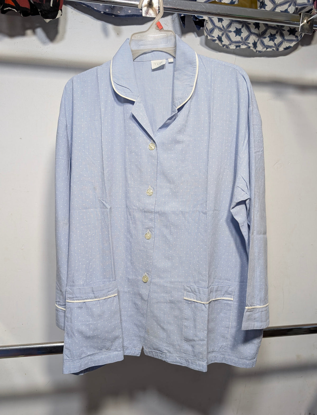 A light blue long-sleeve pajama shirt with white piping trim and gold buttons, hanging on a clear plastic hanger against a white wall.