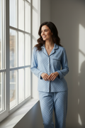 A woman wearing a light blue long-sleeve pajama set with small dark polka dots stands by a large window, looking out with a smile. The room has soft natural light and a neutral gray wall.