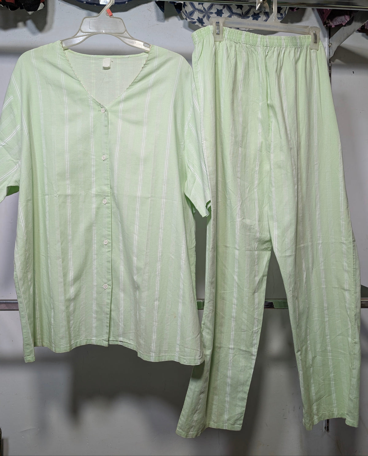 A light green two-piece pajama set consisting of a short-sleeve button-front top and matching pants, hanging on hangers from a clothing rack.