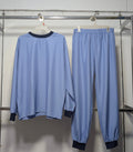 A light blue two-piece tracksuit hanging on a metal rack against a white wall, featuring a long-sleeve top with dark navy cuffs and collar, and matching light blue jogger pants with dark navy ankle cuffs.