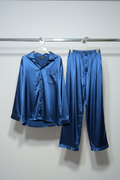 A two-piece satin pajama set in a rich blue color, featuring a long-sleeve button-up shirt and matching wide-leg pants, hanging on white hangers from a metal rail against a plain light gray wall.