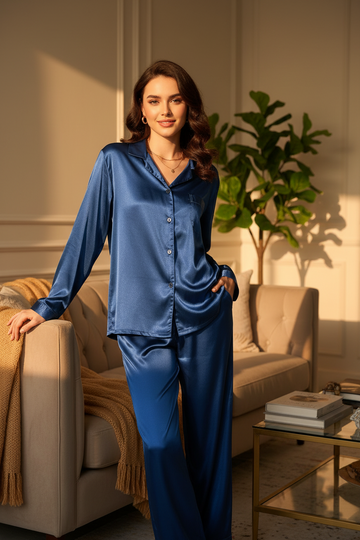 A woman stands in a sunlit living room wearing a satin-like blue pajama set with a collared button-front top and matching wide-leg pants. The scene includes a beige sofa, throw blanket, plant, and small side table.