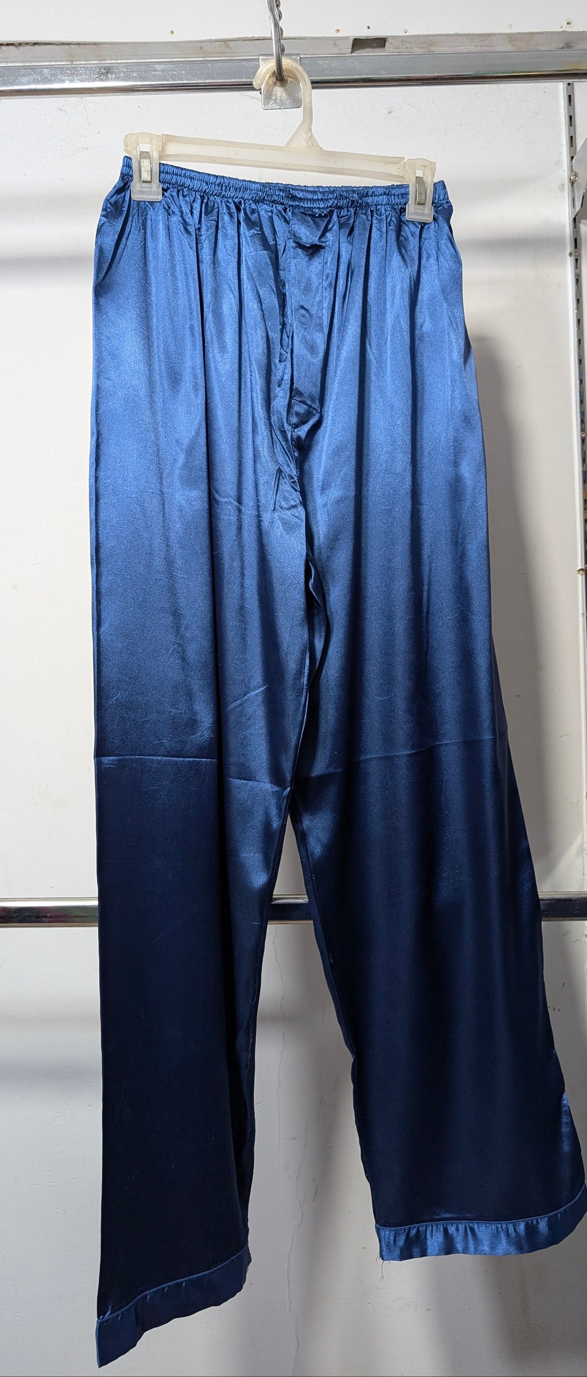 Blue satin cropped pants hanging on a white plastic hanger from a metal clothing rack against a white wall.