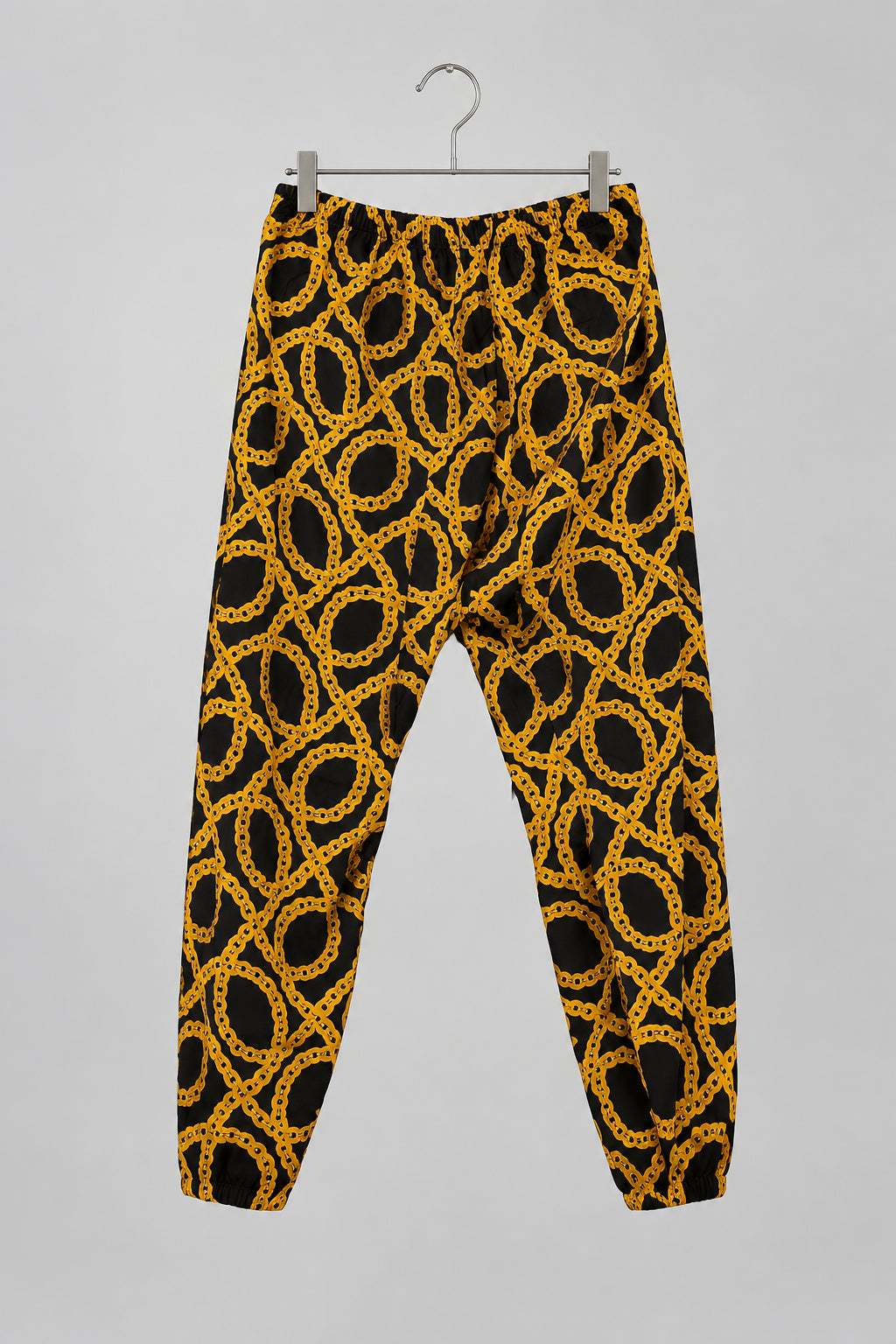 A pair of loose-fitting jogger pants with a bold yellow and black geometric chain-link pattern, hanging on a metal hanger against a white wardrobe door.