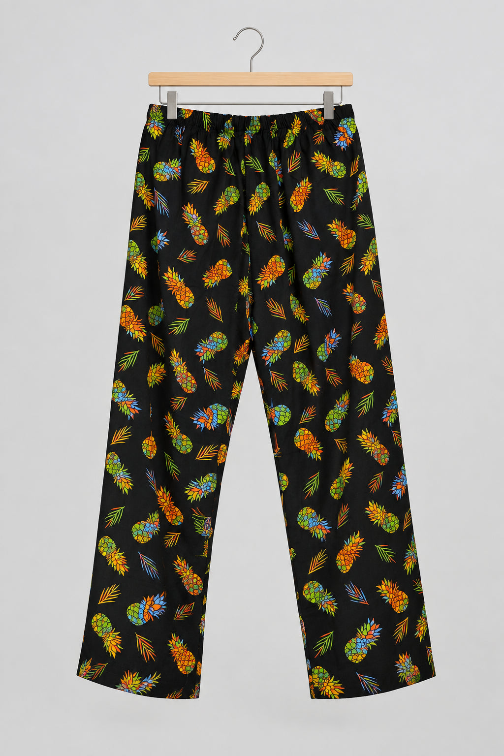 Black pants with a colorful pineapple print, hanging on a metal hanger against a white wardrobe door.