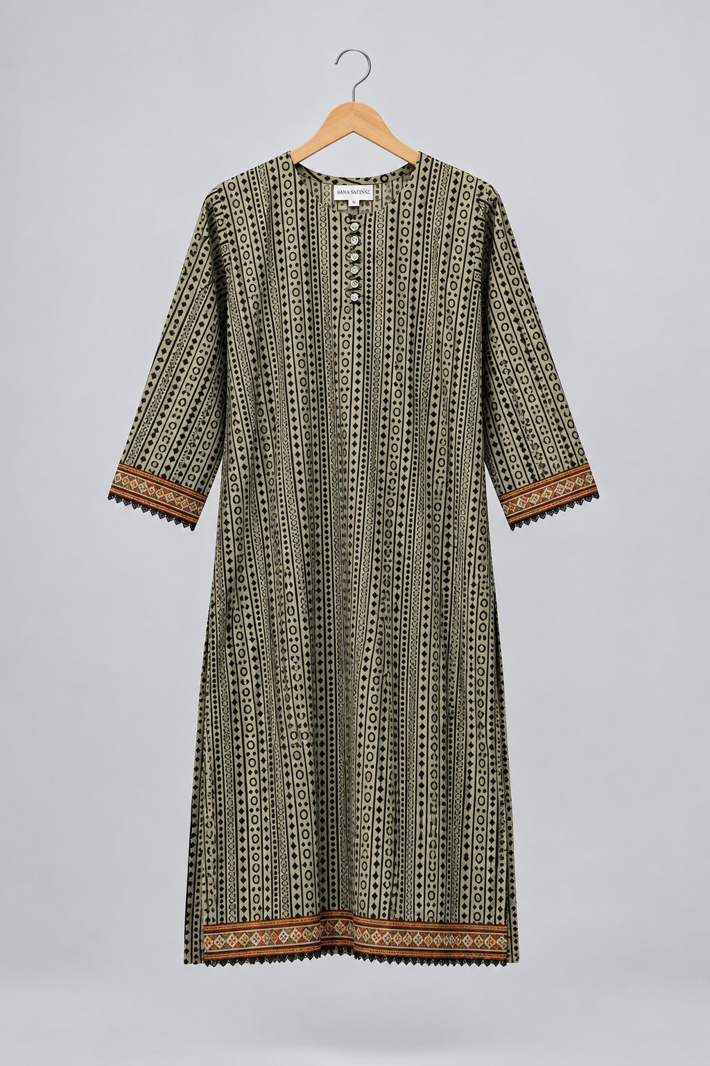 A three-quarter sleeve tunic dress with a vertical geometric pattern in olive green, black, and white, featuring brown and gold patterned borders on the cuffs and hem, hanging on a hanger against a white wardrobe door.