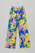 A pair of wide-leg cropped pants with a vibrant floral print, featuring purple, yellow, and green flowers on a blue background, hanging on a metal hanger against a white wardrobe door.