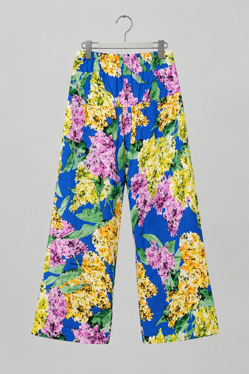 A pair of wide-leg cropped pants with a vibrant floral print, featuring purple, yellow, and green flowers on a blue background, hanging on a metal hanger against a white wardrobe door.