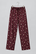 A pair of loose-fitting, long pajama pants in a dark burgundy fabric with a small white and pink floral print, hanging on a metal hanger against a white wardrobe door.