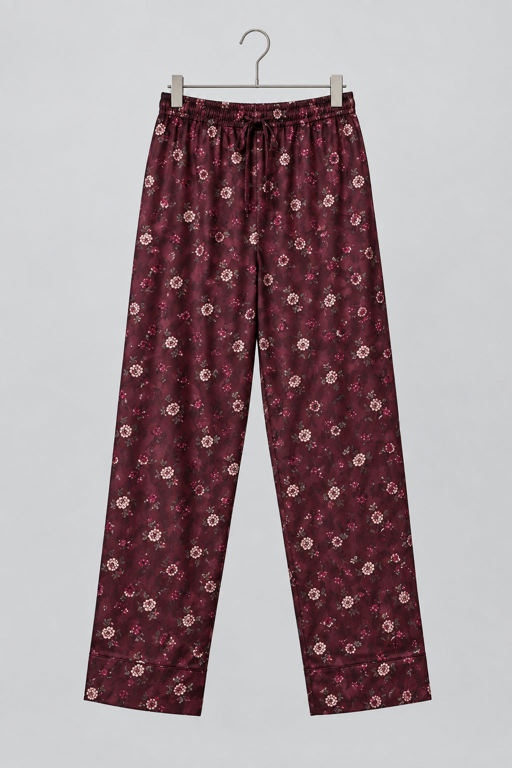 A pair of loose-fitting, long pajama pants in a dark burgundy fabric with a small white and pink floral print, hanging on a metal hanger against a white wardrobe door.