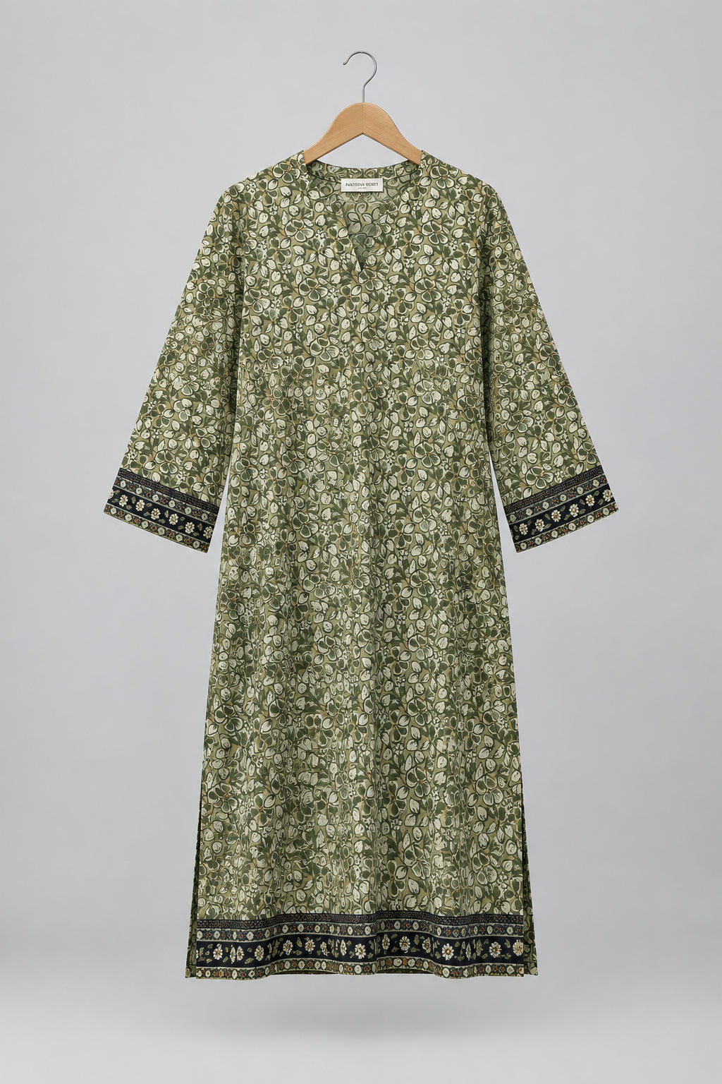A long-sleeved tunic dress with a dense green and beige floral print, featuring dark patterned trim on the cuffs and hem, hanging on a hanger against a white wardrobe door.