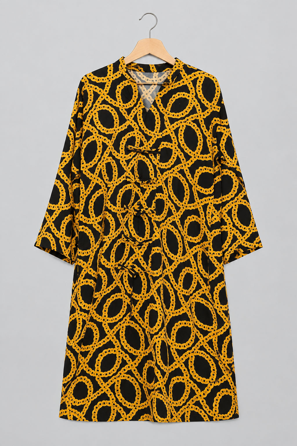 A black and yellow patterned tunic top with a chain-link design, hanging on a metal hanger against a white wardrobe door, with a hand holding the hem on the left.