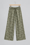 A pair of loose, cropped pants with an all-over green and beige floral or leafy pattern, hanging on a metal hanger against a white wardrobe door.