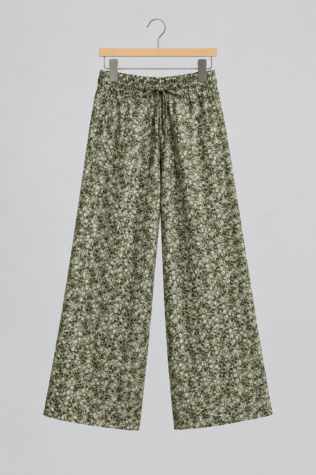 A pair of loose, cropped pants with an all-over green and beige floral or leafy pattern, hanging on a metal hanger against a white wardrobe door.