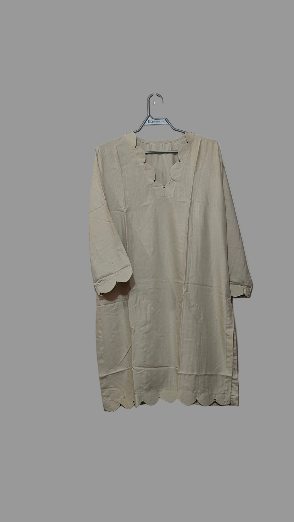 A light beige linen tunic dress with scalloped edges, hanging on a metal hanger against a white wardrobe door.