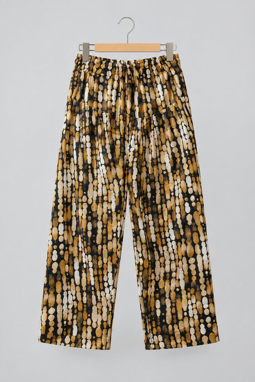 A pair of loose, cropped pants with an abstract black, brown, and white spotted pattern, hanging on a metal hanger against a white wardrobe door.