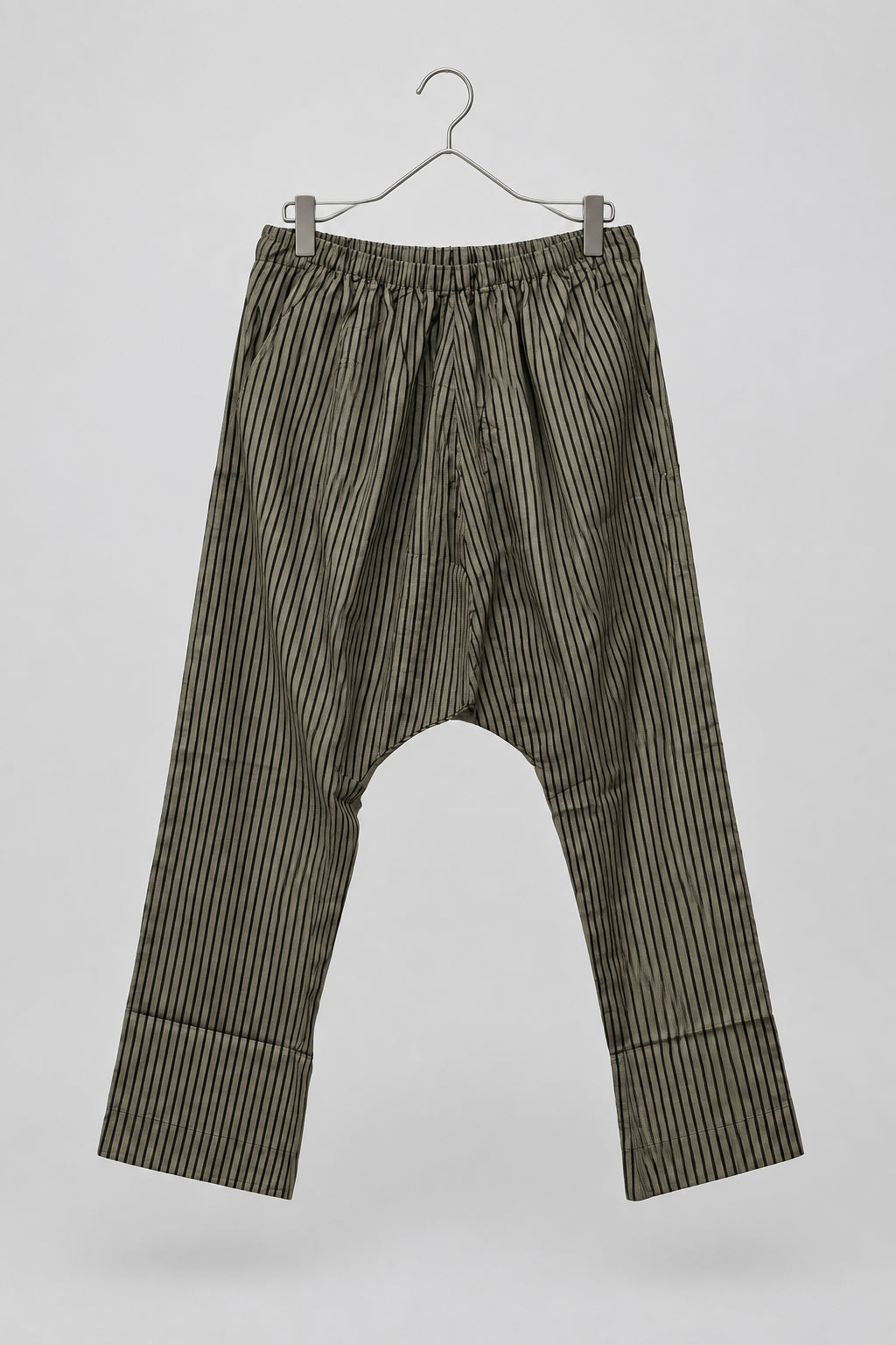 A pair of loose-fitting striped pants hanging on a metal hanger against a white wardrobe door. The pants have vertical black and olive-brown stripes and a gathered elastic waistband.