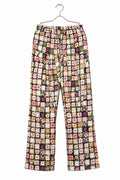 A pair of patterned cropped pants hanging on a metal hanger against a white wardrobe door. The pants feature a busy patchwork-style print with small floral and geometric motifs in pink, purple, green, yellow, and beige tones.